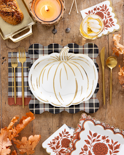A styled autumn table setting featuring a white scalloped plate with a gold pumpkin outline, a black and white plaid napkin, gold flatware, and coordinating patterned napkins on a rustic wooden table.