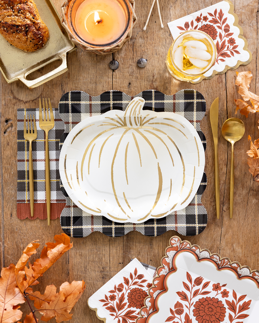 A styled autumn table setting featuring a white scalloped plate with a gold pumpkin outline, a black and white plaid napkin, gold flatware, and coordinating patterned napkins on a rustic wooden table.