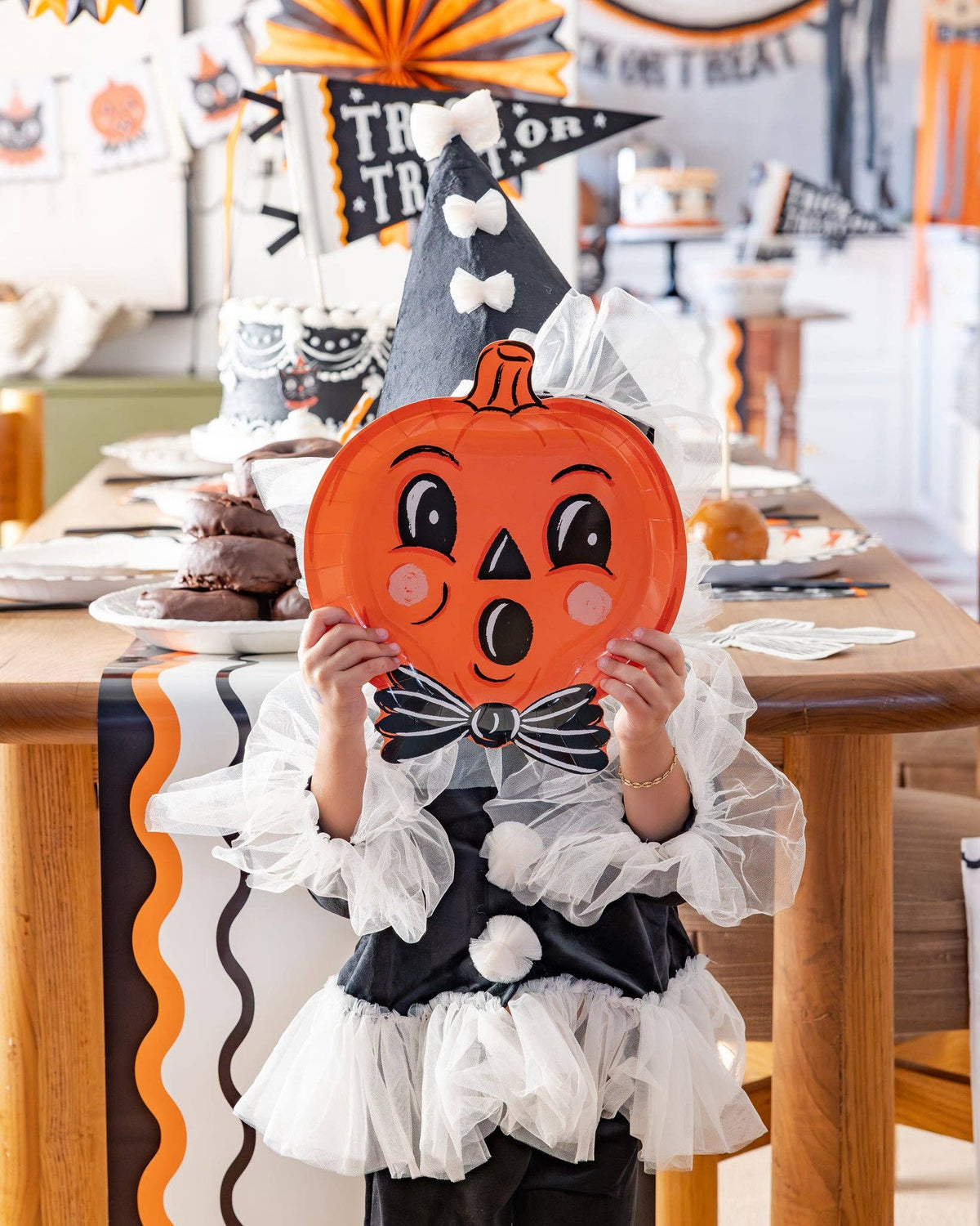 Child in Halloween costume holding a pumpkin-shaped plate with a festive table setting in the background.
