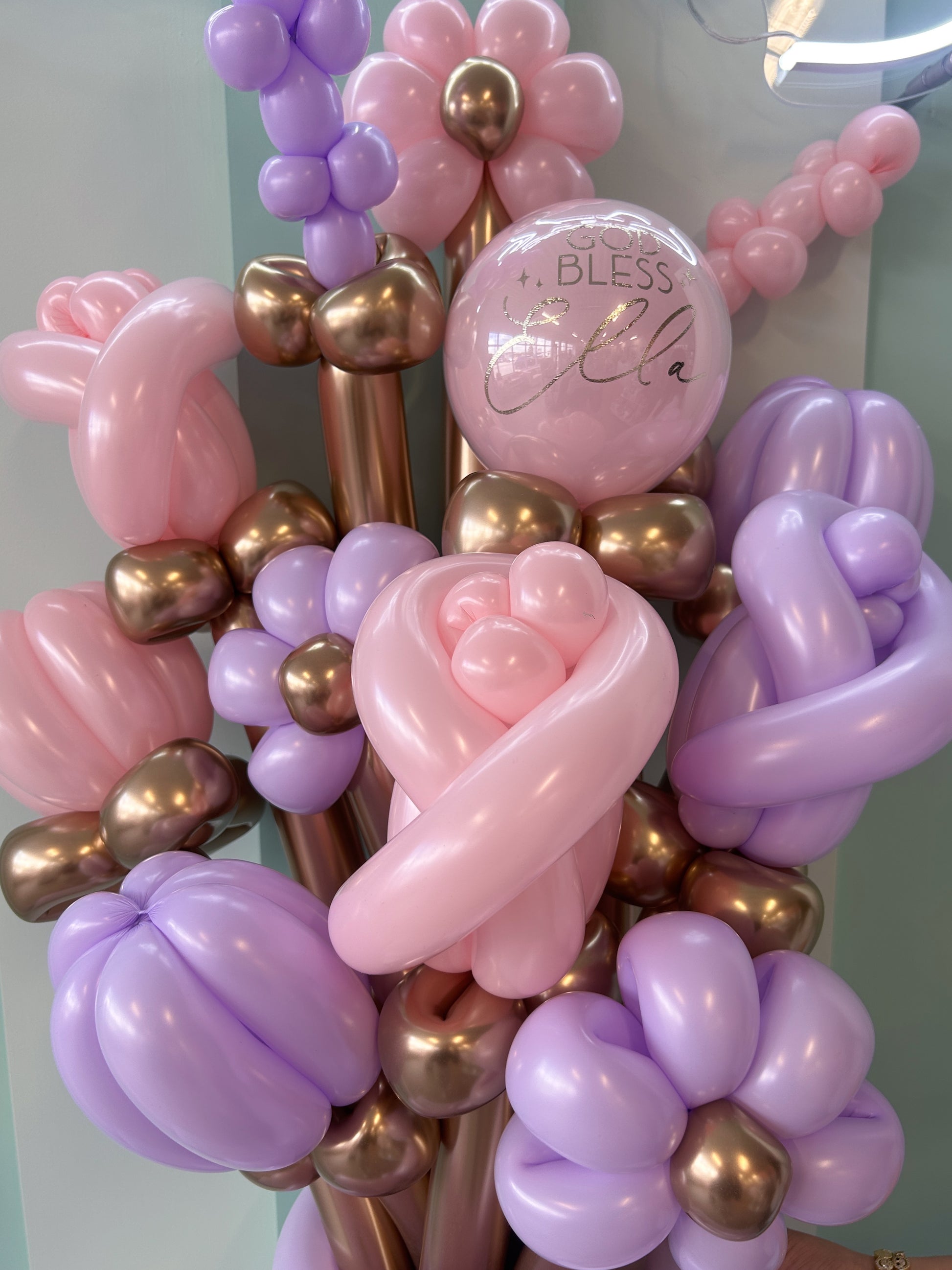 Balloon bouquet with pink, purple, and gold balloons, featuring a 'Bless' balloon in the center.