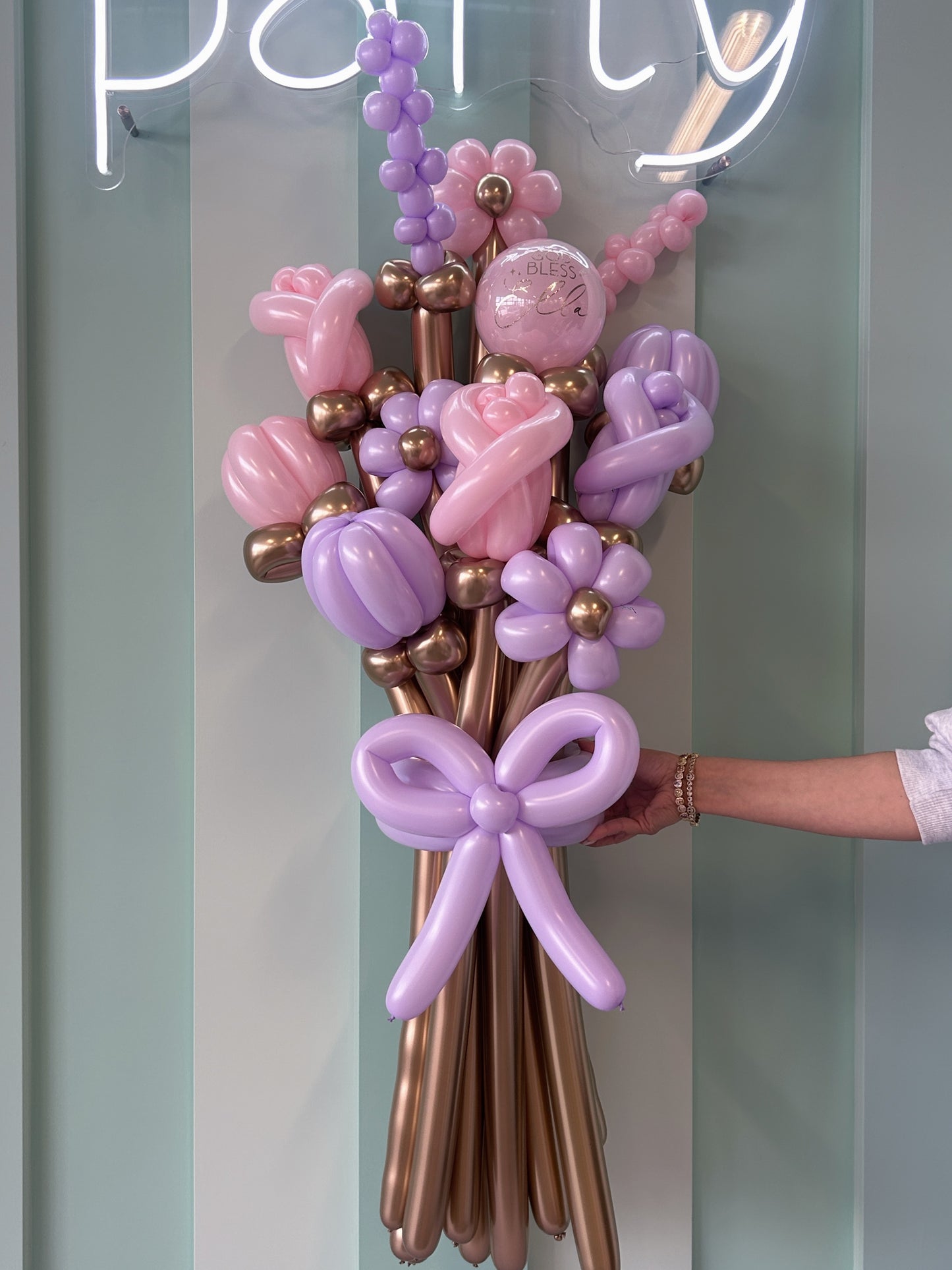 Balloon bouquet with pink, purple, and gold balloons held by a person in front of a neon 'party' sign.