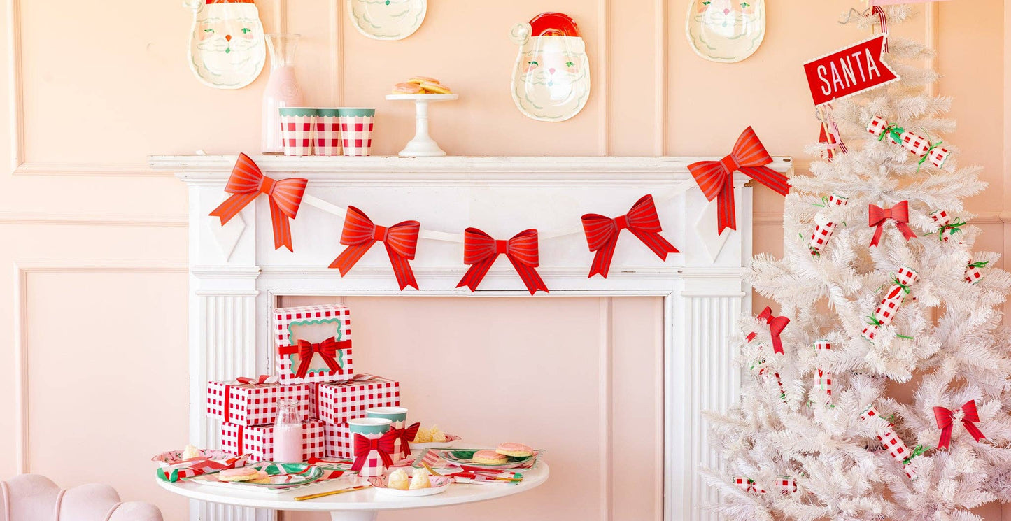 Red bow banner hanging across white fireplace mantel with pink holiday décor, Christmas setup inspo.