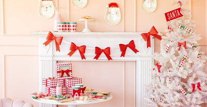 Red bow banner hanging across white fireplace mantel with pink holiday décor, Christmas setup inspo.