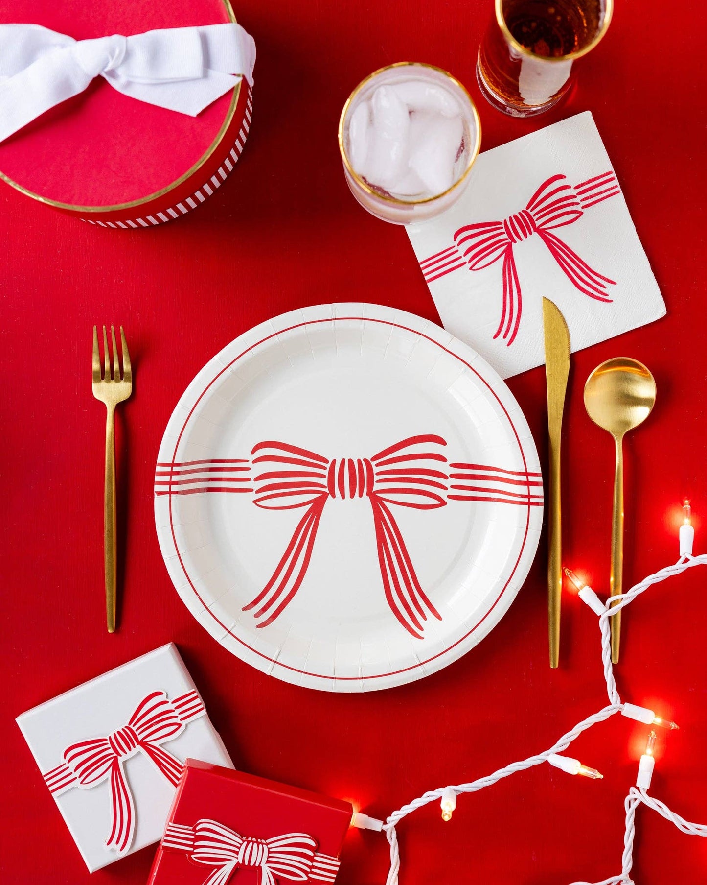 Dining table setting with red and white bow-themed plates, gold cutlery, and small gift boxes on a red background.