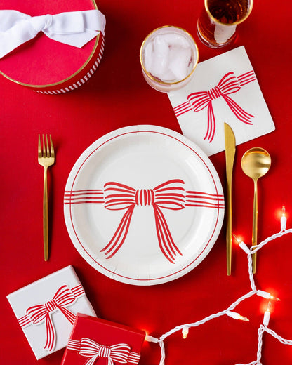 Dining table setting with red and white bow-themed plates, gold cutlery, and small gift boxes on a red background.