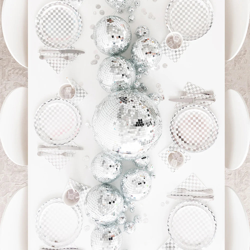 Top-down view of a party tablescape featuring silver checkered plates, napkins, and oversized disco ball centerpiece — ideal for a sparkling, retro-inspired event or New Year's Eve dinner.
