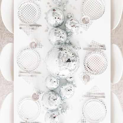 Top-down view of a party tablescape featuring silver checkered plates, napkins, and oversized disco ball centerpiece — ideal for a sparkling, retro-inspired event or New Year's Eve dinner.