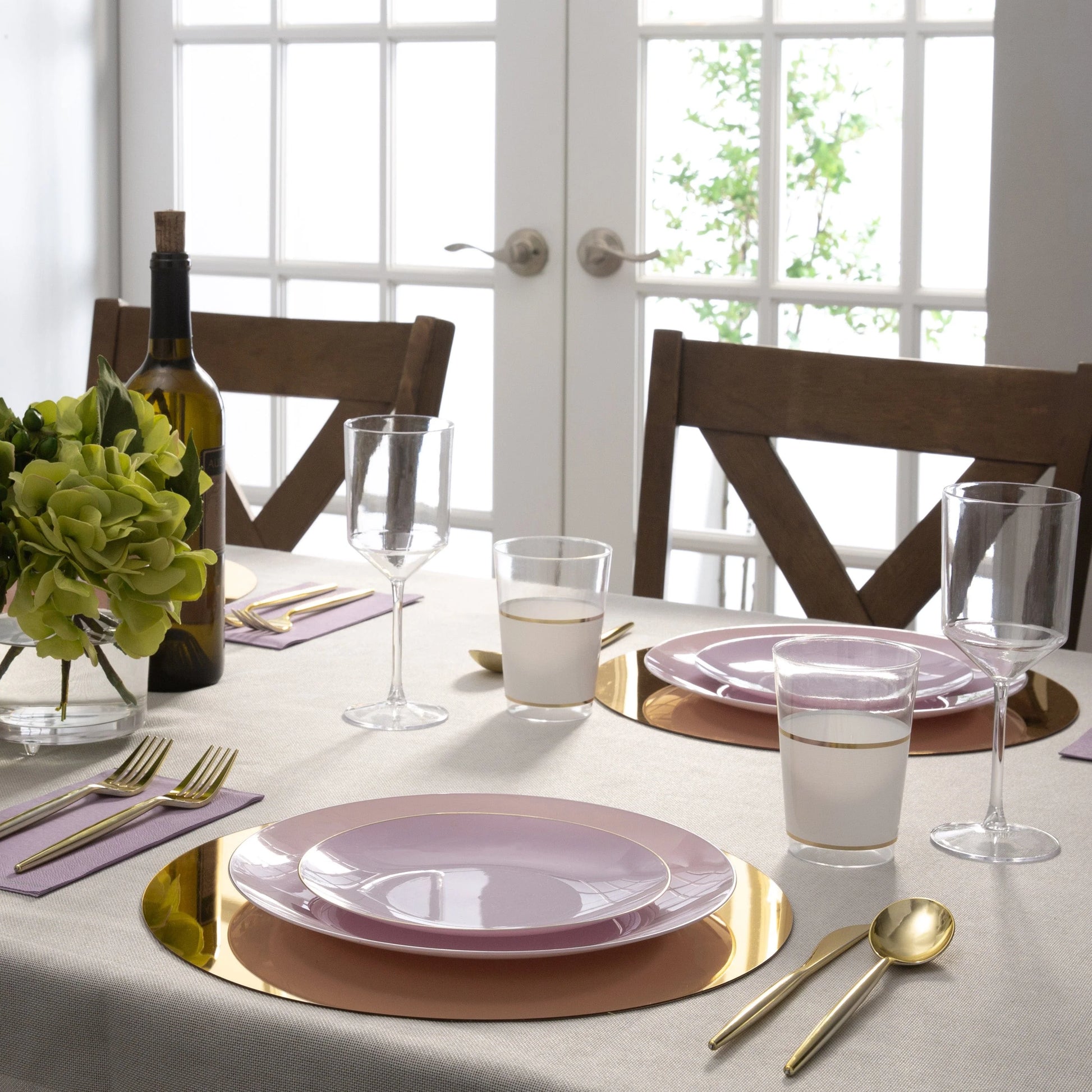 Dining table set with pink plates, gold cutlery, and glasses in a bright room.
