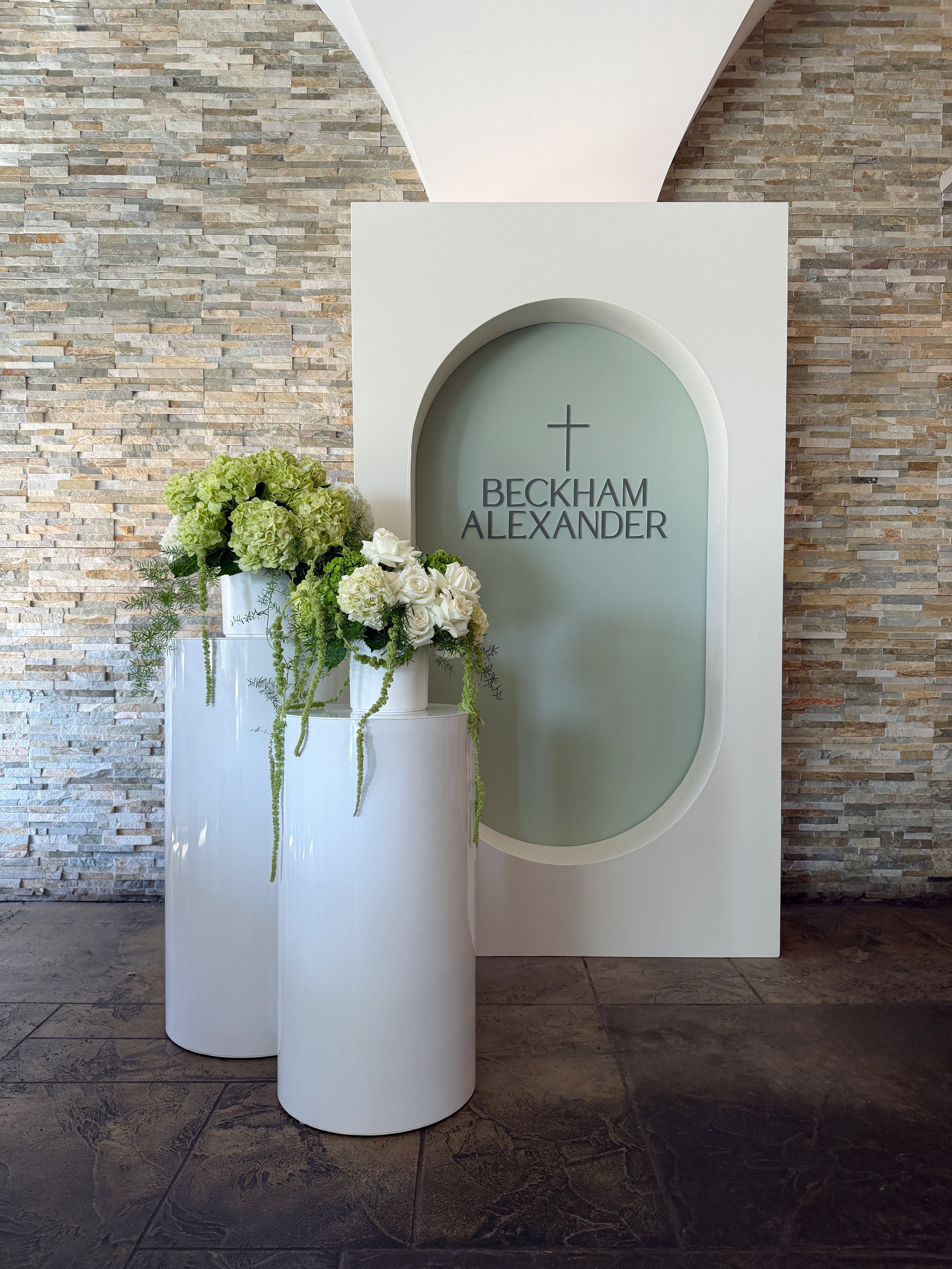 Sacrament backdrop with sage oval panel, cross detail and two white plinths with florals available at our Toronto party shop
