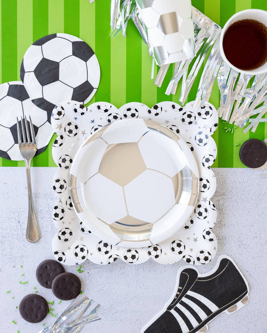 Soccer-themed table setting with plates, cutlery, and decorative items on a green and white striped background.