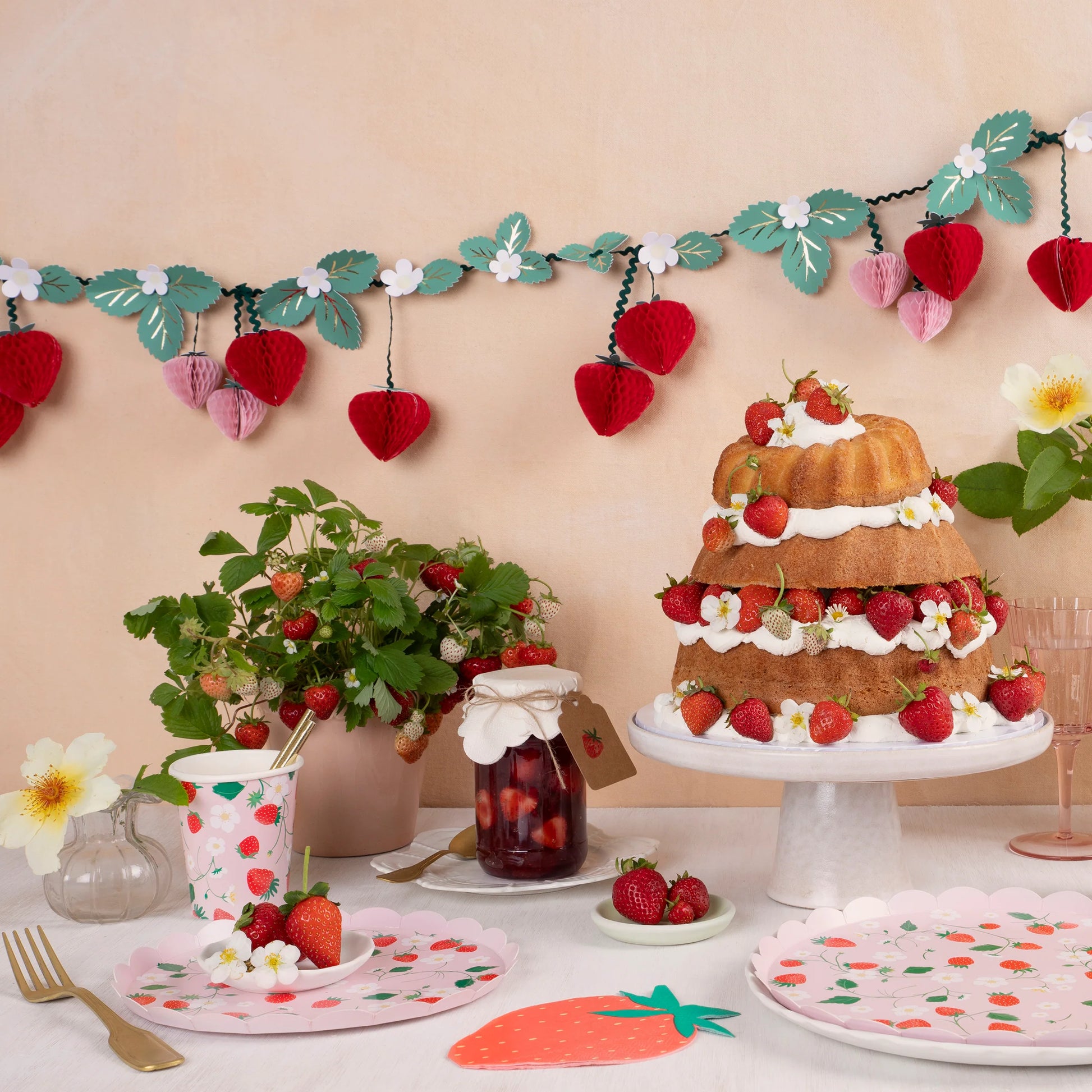 Strawberry-themed party setup featuring tiered cake, floral decor, and hanging garland