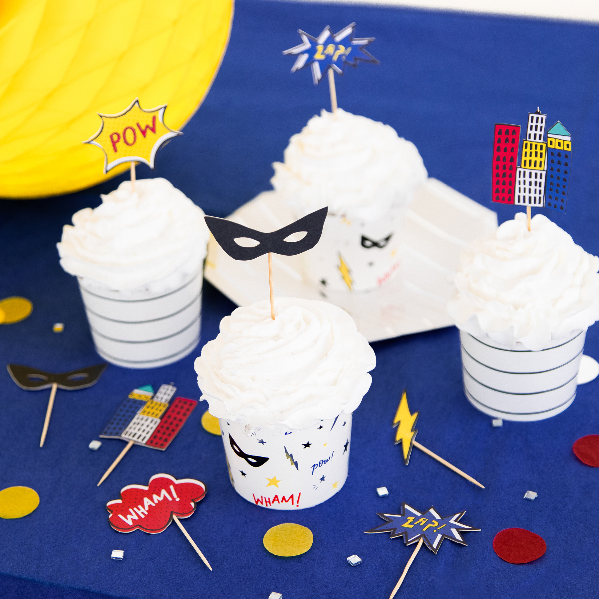 Superhero-themed party setup with cupcakes, masks, and props on a blue tablecloth.