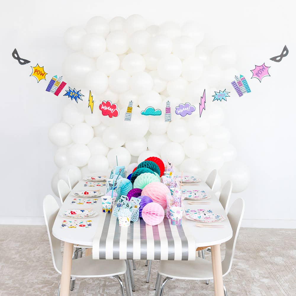 A children's party setup with a white table covered in a gray-and-white striped tablecloth, colorful paper fans, plates, cups, and a superhero-themed balloon garland hanging against a white balloon wall.