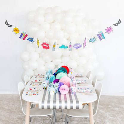 A children's party setup with a white table covered in a gray-and-white striped tablecloth, colorful paper fans, plates, cups, and a superhero-themed balloon garland hanging against a white balloon wall.