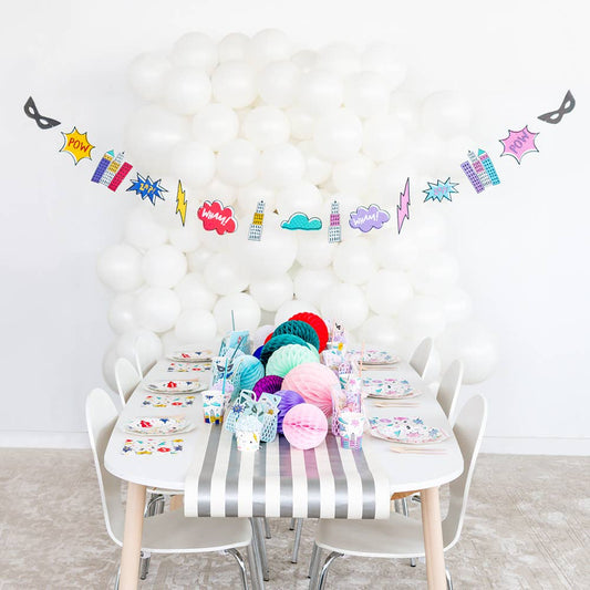 A children's party setup with a white table covered in a gray-and-white striped tablecloth, colorful paper fans, plates, cups, and a superhero-themed balloon garland hanging against a white balloon wall.
