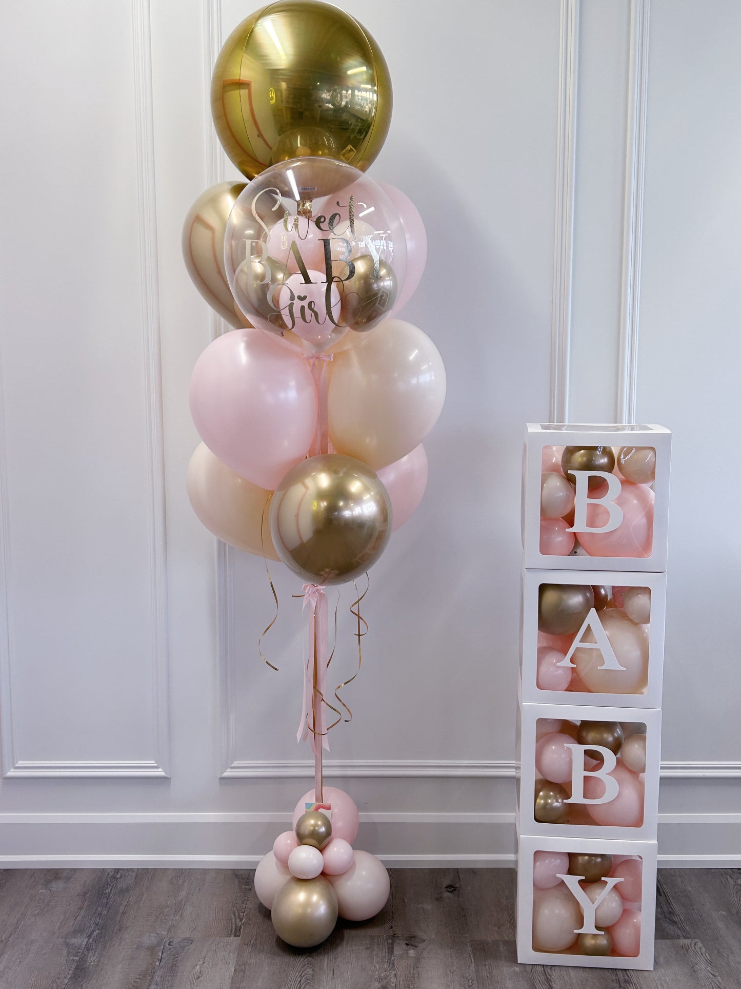 A decorative balloon arrangement in blush pink, metallic gold, and clear balloons with 'sweet baby girl" lettering, standing next to a stack of white blocks displaying the letters B, A, B, Y.