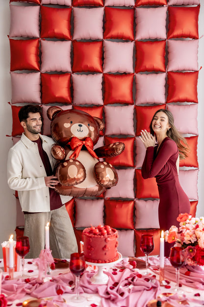 Couple holding large brown teddy bear foil balloon in front of red and pink balloon wall, available from Confetti My Party Toronto with Canada-wide shipping.