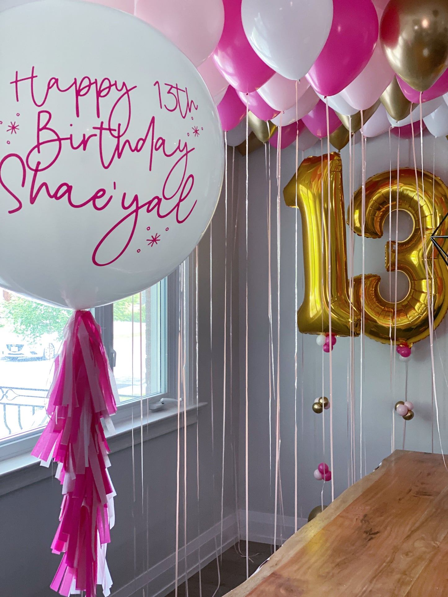 Pink, white, and gold birthday balloon decorations in a room, featuring a large white balloon with 'Happy 13th Birthday' in pink script, a gold foil number 13 balloon, and pink and white ceiling balloons.