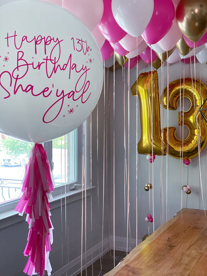 Pink, white, and gold birthday balloon decorations in a room, featuring a large white balloon with 'Happy 13th Birthday' in pink script, a gold foil number 13 balloon, and pink and white ceiling balloons.
