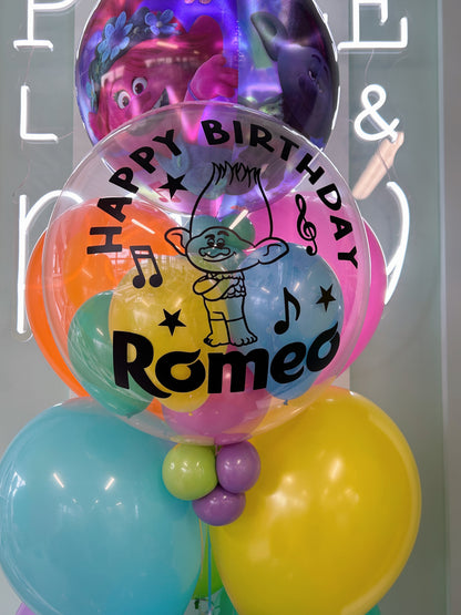 Close-up of personalized Trolls birthday bubble balloon reading Happy Birthday Romeo with Branch character graphic, colourful mini balloons and Trolls foil balloon.