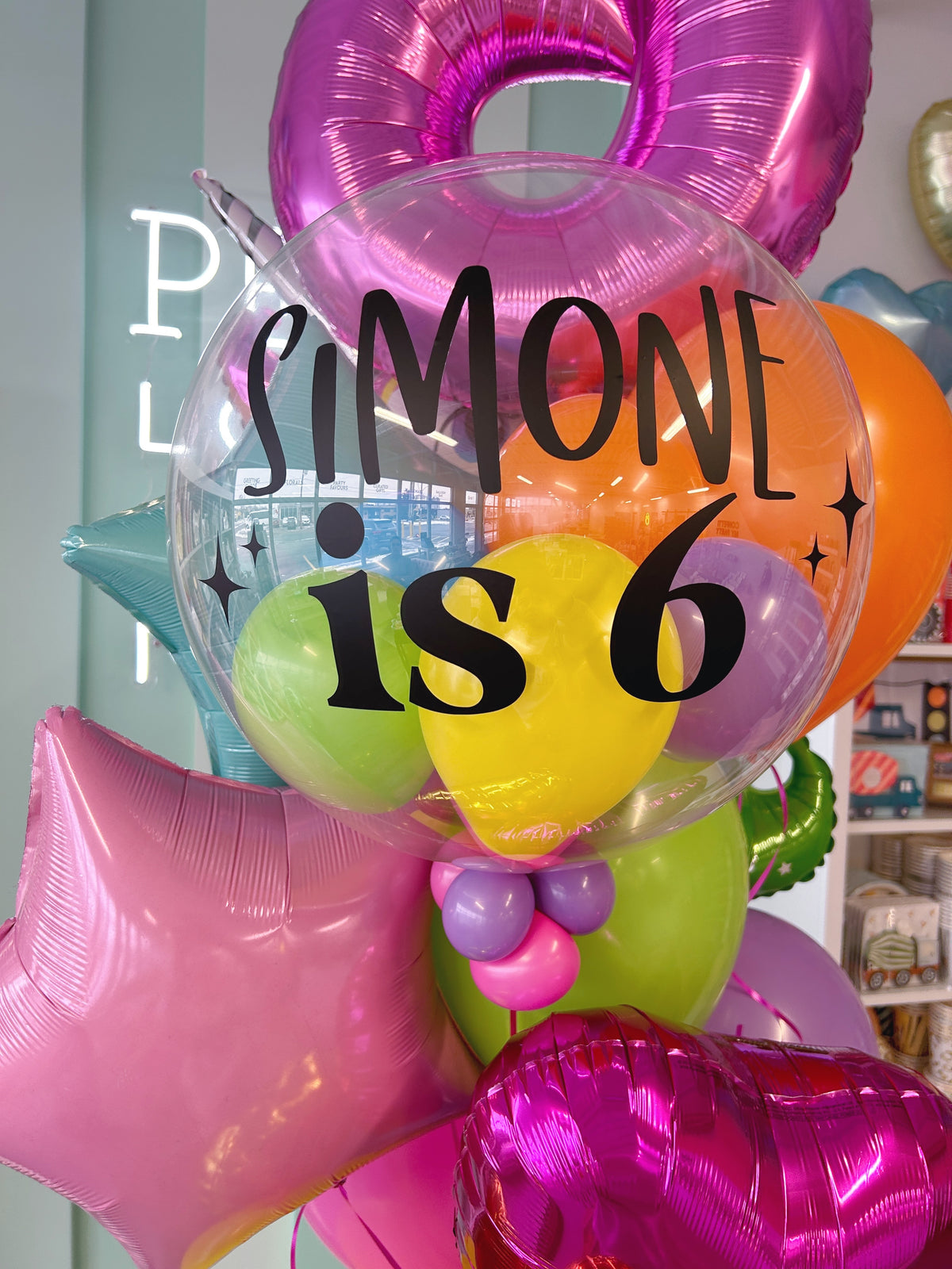 Personalized clear bubble balloon reading Simone is 6, surrounded by bright rainbow latex balloons, pink foil stars and unicorn birthday balloons.