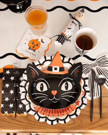Halloween-themed table setting with black cat plate, orange and black napkins, and drinks on a wooden table.