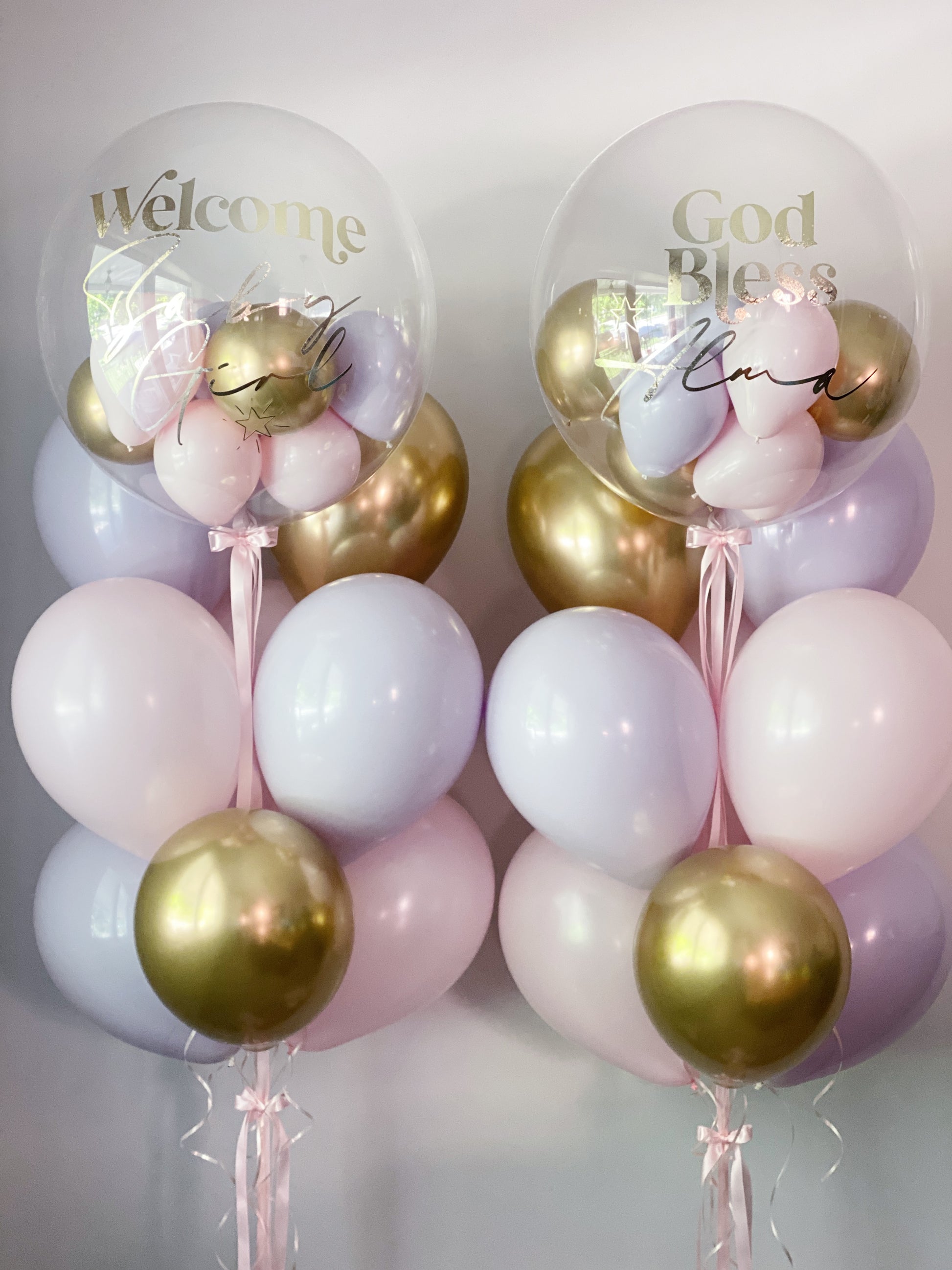 Two balloon arrangements with clear round balloons printed with 'Welcome Baby' and 'God Bless Baby,' filled with pastel pink, lavender, and metallic gold balloons, tied with pink ribbons against a light gray background.