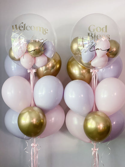 Two balloon arrangements with clear round balloons printed with 'Welcome Baby' and 'God Bless Baby,' filled with pastel pink, lavender, and metallic gold balloons, tied with pink ribbons against a light gray background.