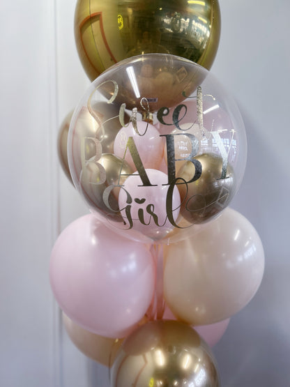 A decorative balloon garland featuring a large clear round balloon with 'Sweet Baby Girl' printed in metallic script and block letters, surrounded by pastel pink, cream, and metallic gold balloons against a light gray wall.