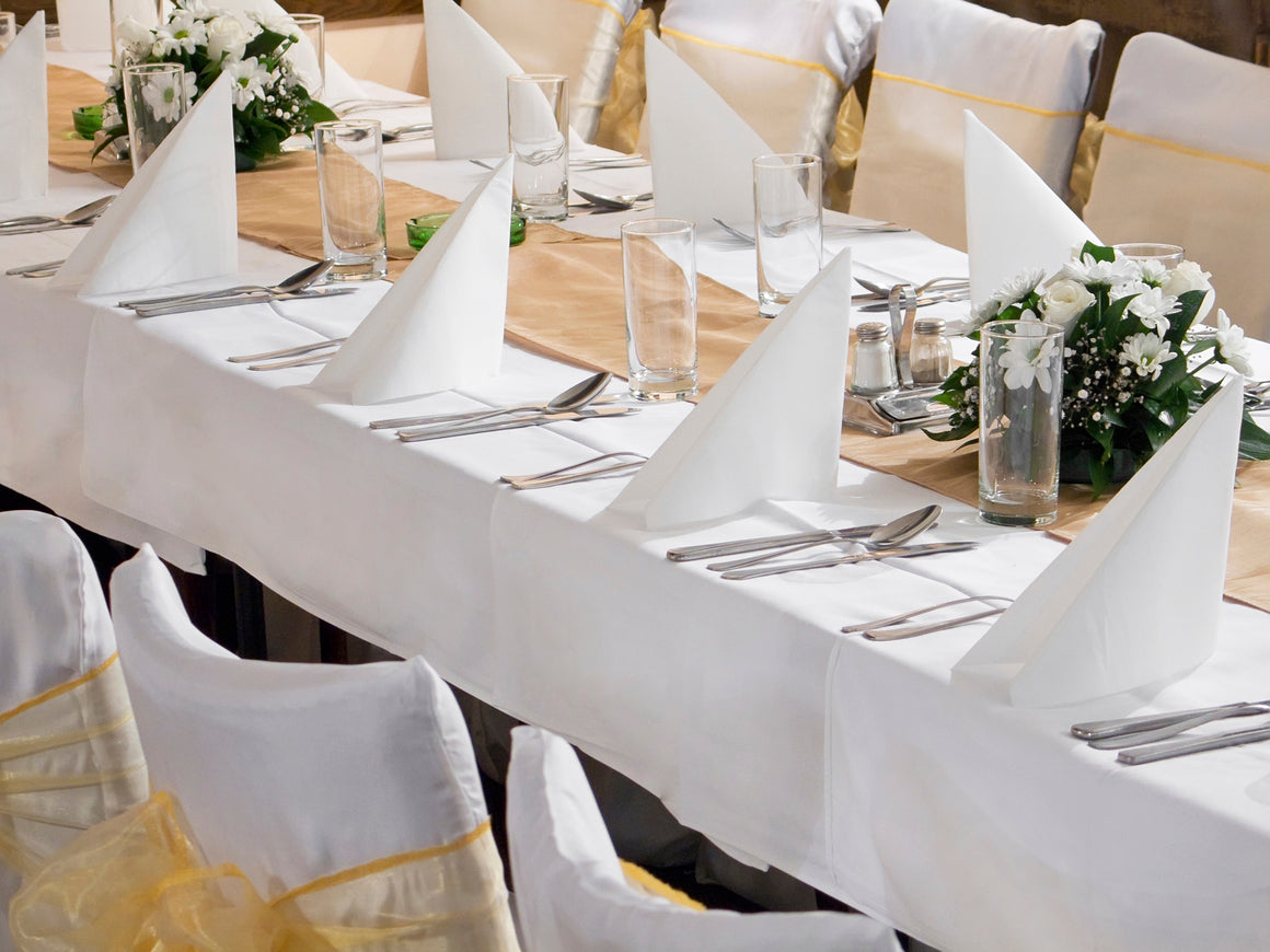 Elegant banquet table setup with white rectangular linen tablecloths, folded napkins, glassware, silver cutlery, and white floral centerpieces. Perfect for weddings, receptions, or formal events.