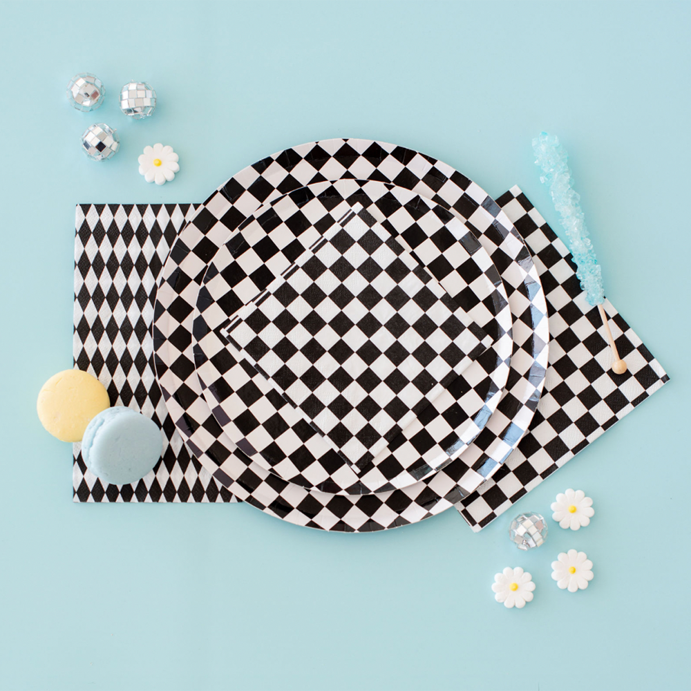 Flat lay of black and white checkered party plates and napkins styled with pastel macarons, mini disco balls, daisies, and blue rock candy on a light blue background.